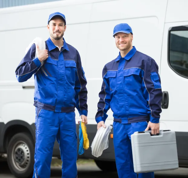 about-1 Two men in blue uniforms stand by a white van, holding cables and a toolbox, smiling at the camera.
