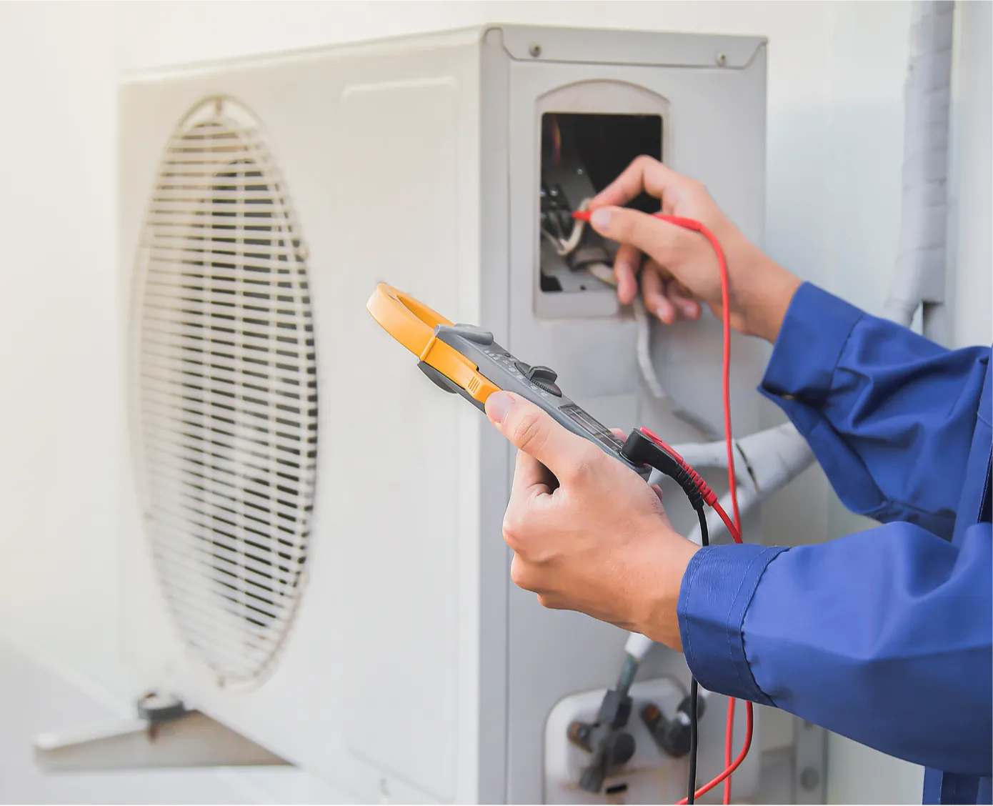 ac-repair-1 Technician in blue uniform testing an air conditioner unit with an electric multimeter.