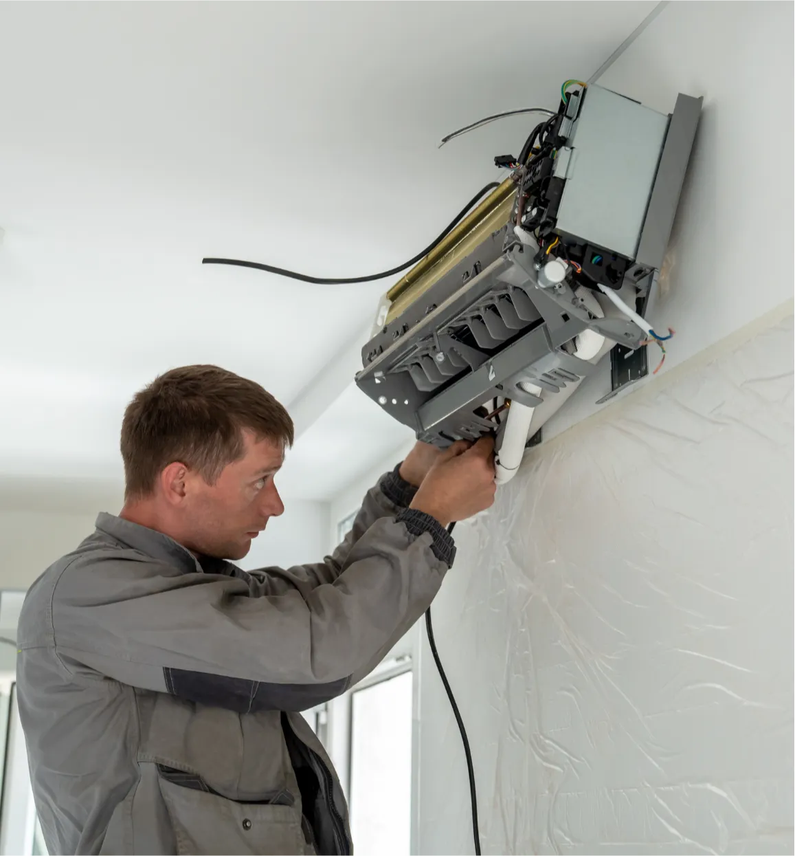 ac-repair-3 A technician repairing an air conditioner unit mounted on a white wall indoors.