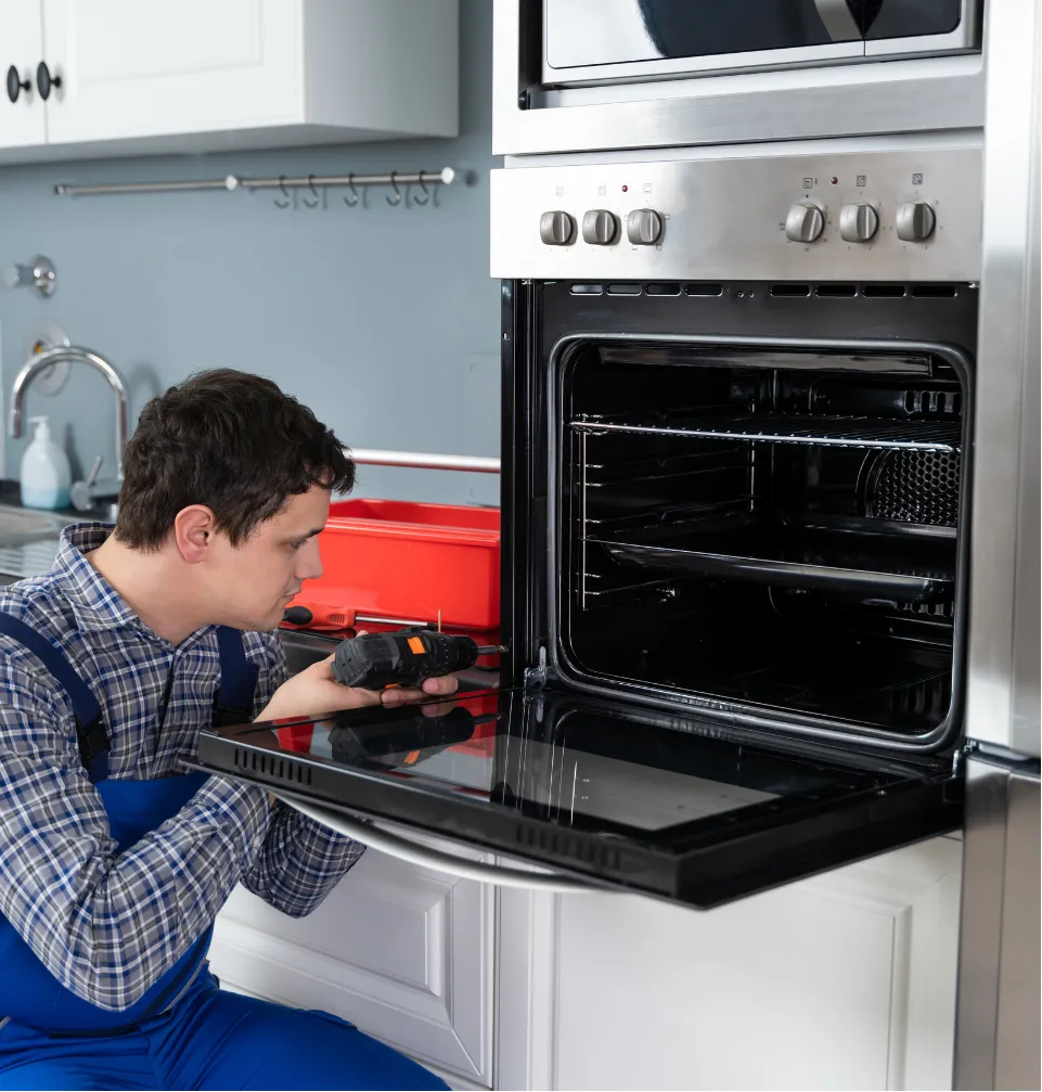 appliances-2 A repairman in blue overalls kneels, using a screwdriver to fix an open kitchen oven.