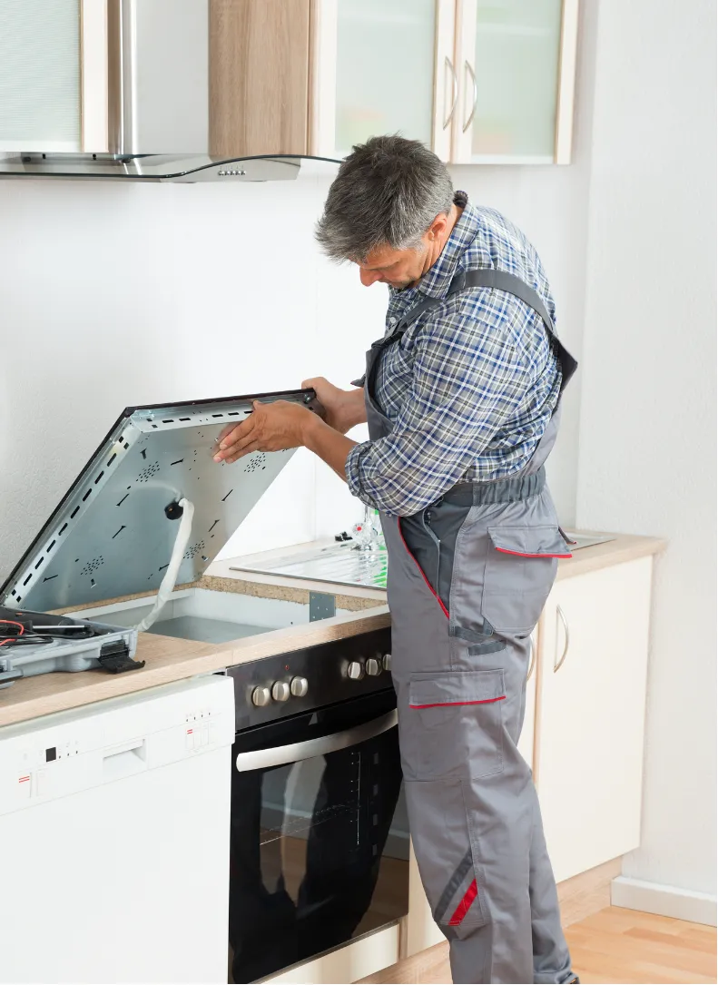 appliances-3 A repairman in overalls fixes a stovetop in a modern kitchen.