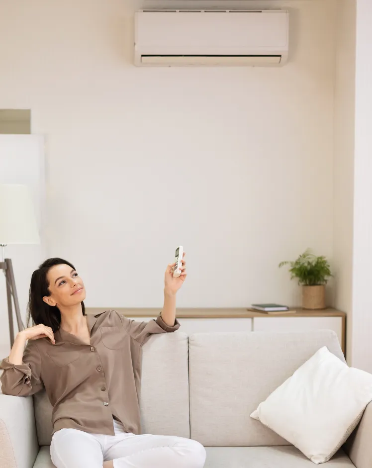 comfort-2 Woman sitting on a couch using a remote to operate an air conditioner mounted on the wall above her.