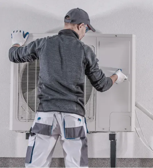 heating-install-1 Technician in uniform servicing an outdoor air conditioning unit mounted on a wall.