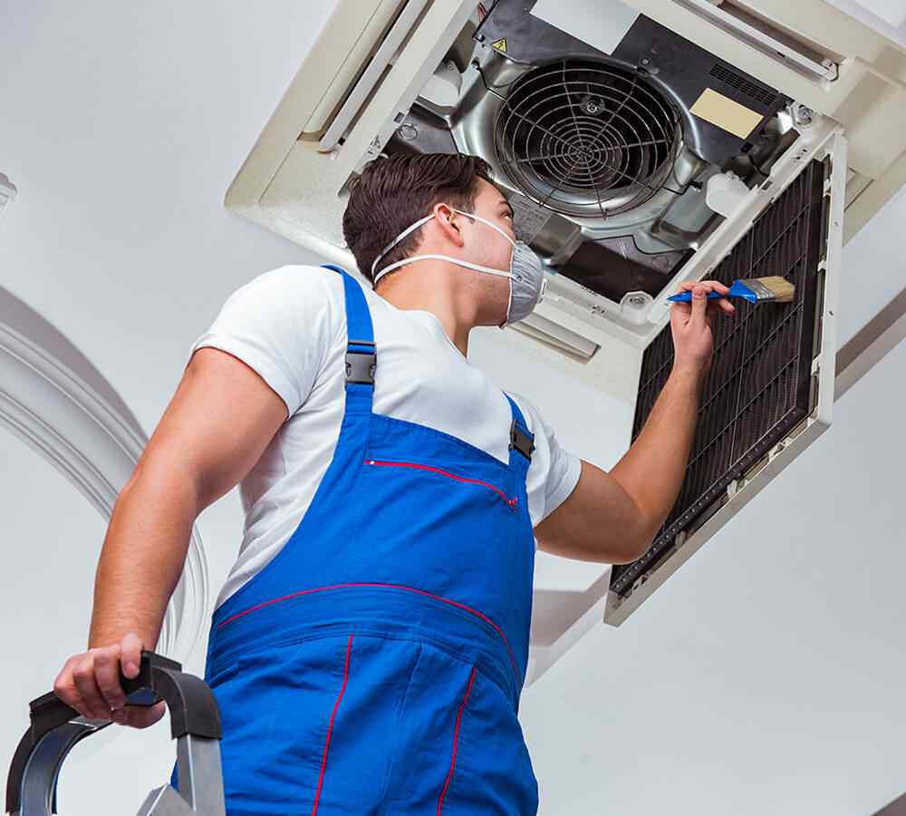inner_12-1 Technician in blue overalls cleaning an air conditioning unit while standing on a ladder.