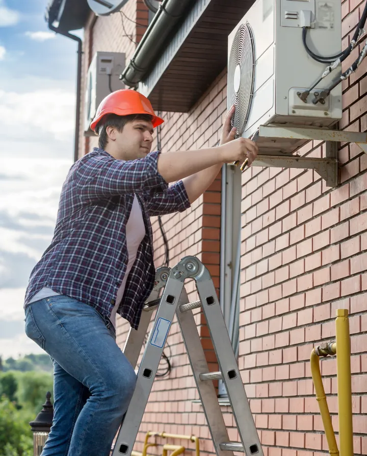 why-choose-2 Man in hard hat on ladder repairing an outdoor air conditioning unit attached to a brick wall.