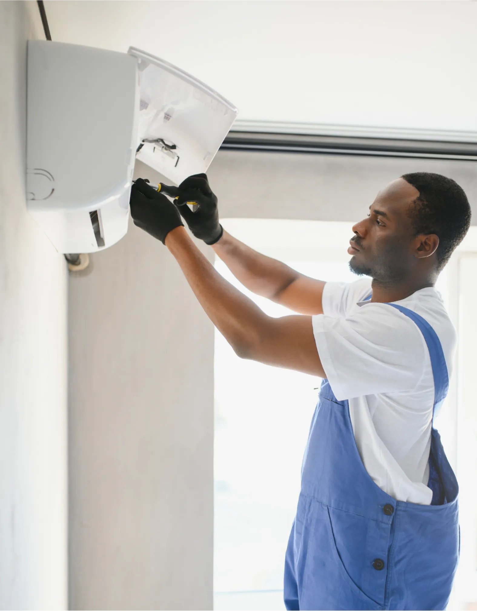 ac-inst-w A technician in blue overalls repairs an air conditioner mounted on a wall indoors.