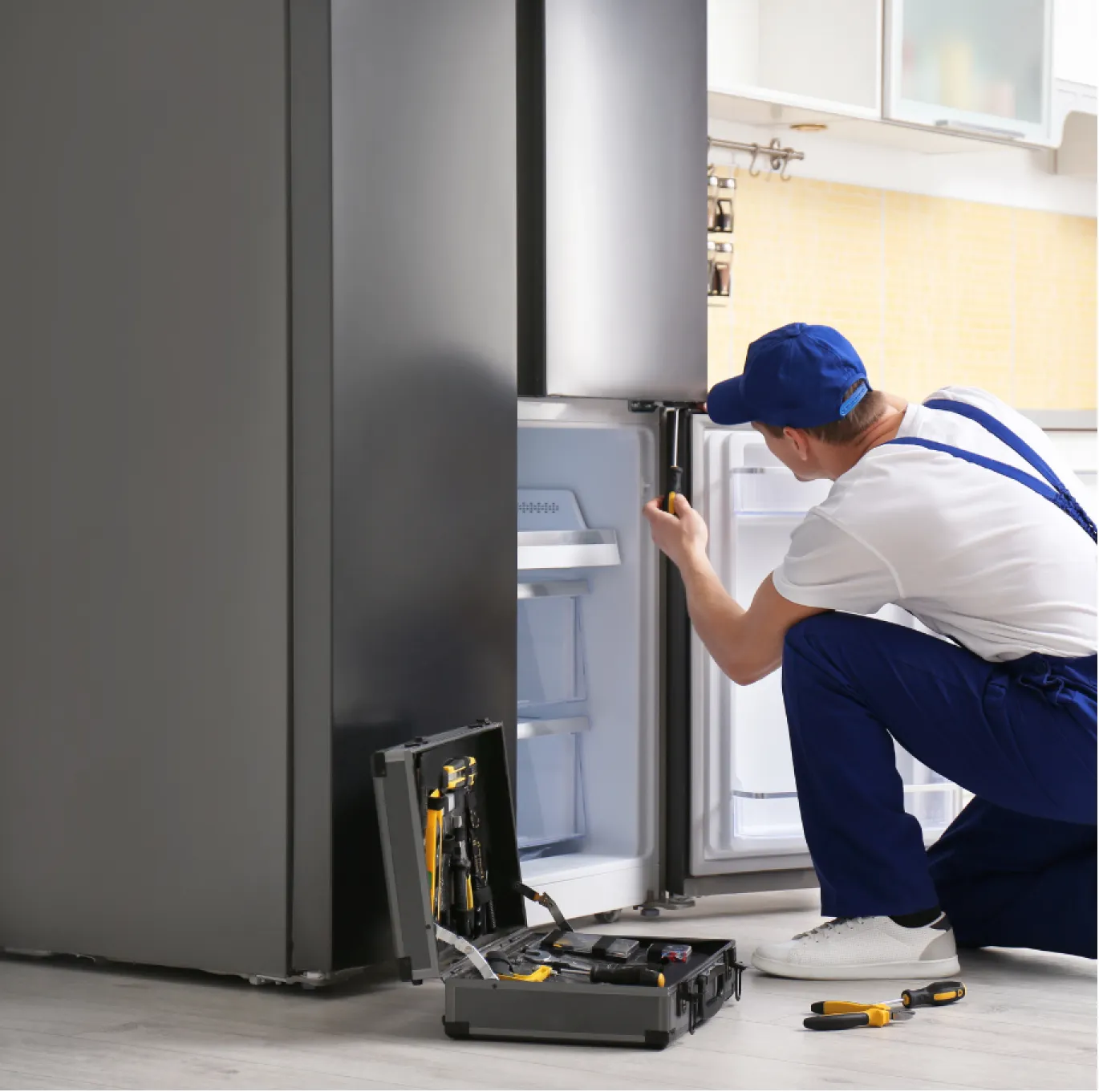 appliance-1 A repair technician in uniform fixes a refrigerator with tools on the floor beside him.