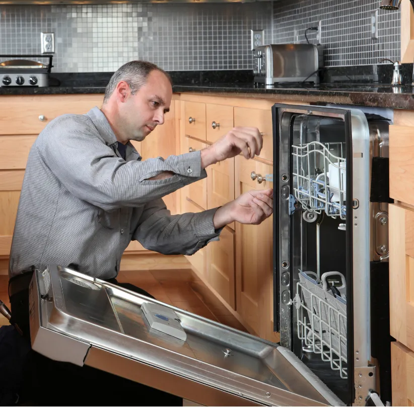 Man repairing an open dishwasher in a modern kitchen with wooden cabinets and tiled backsplash.