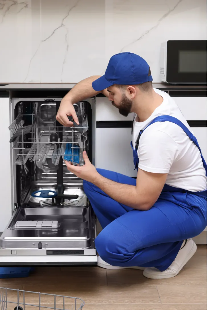 A repairman in blue overalls fixes an open dishwasher in a modern kitchen.