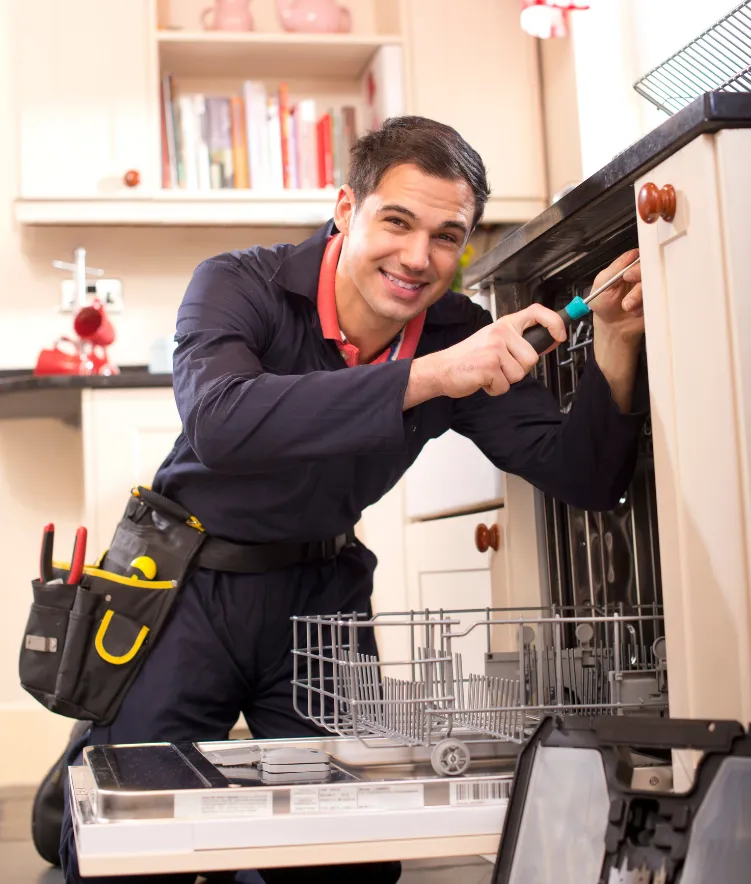 Smiling repairman in uniform fixing a dishwasher with a screwdriver in a modern kitchen.