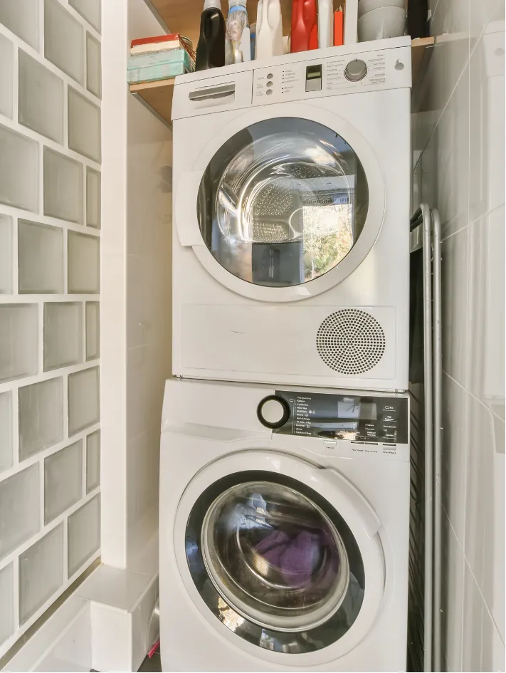 Stacked washing machine and dryer in a small laundry space with cleaning products on a shelf above.