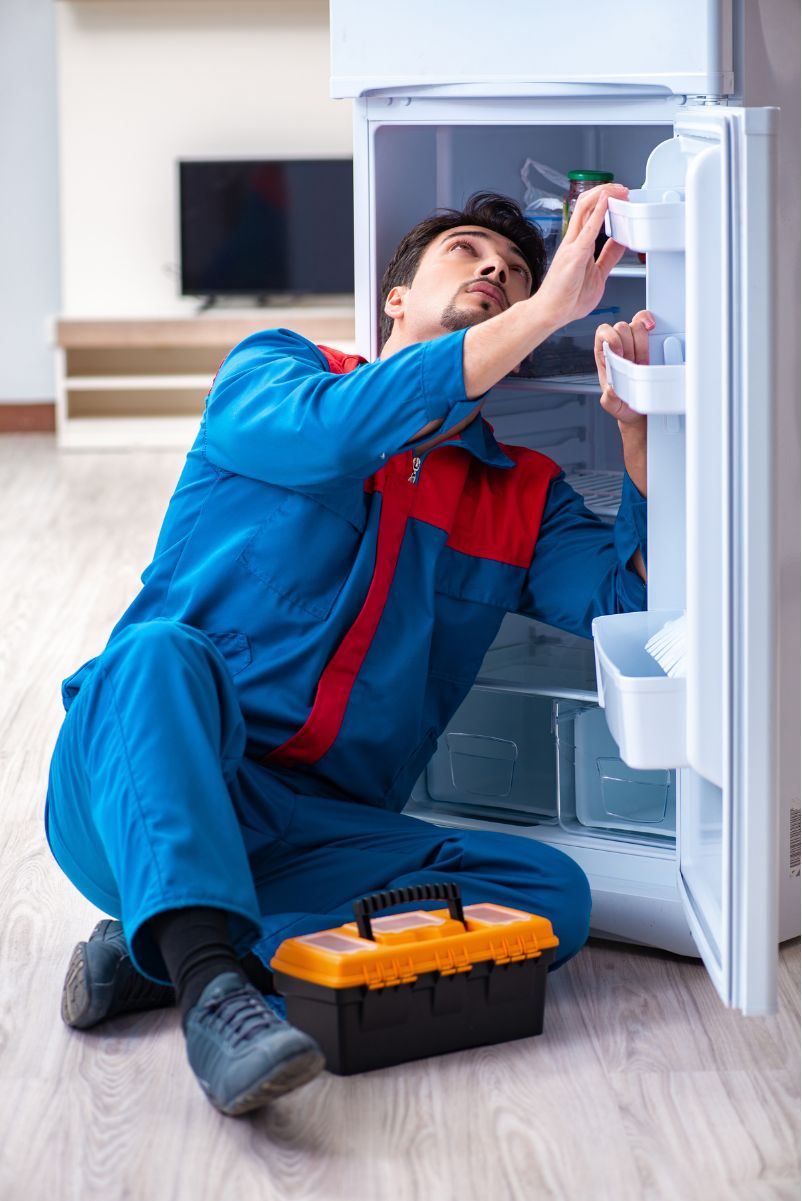 A repairman in blue overalls fixing a refrigerator with a toolbox on the floor beside him.