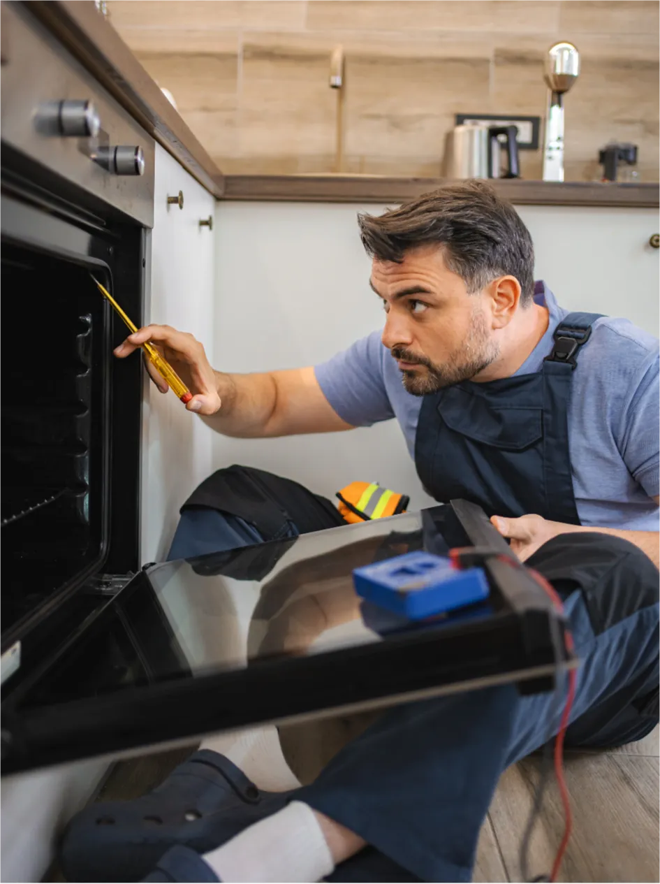 Man in work overalls checking a kitchen oven with a multimeter and yellow measuring tape.