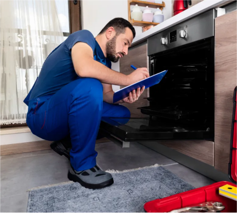 Man in blue uniform inspects an open oven and takes notes on a clipboard in a modern kitchen.