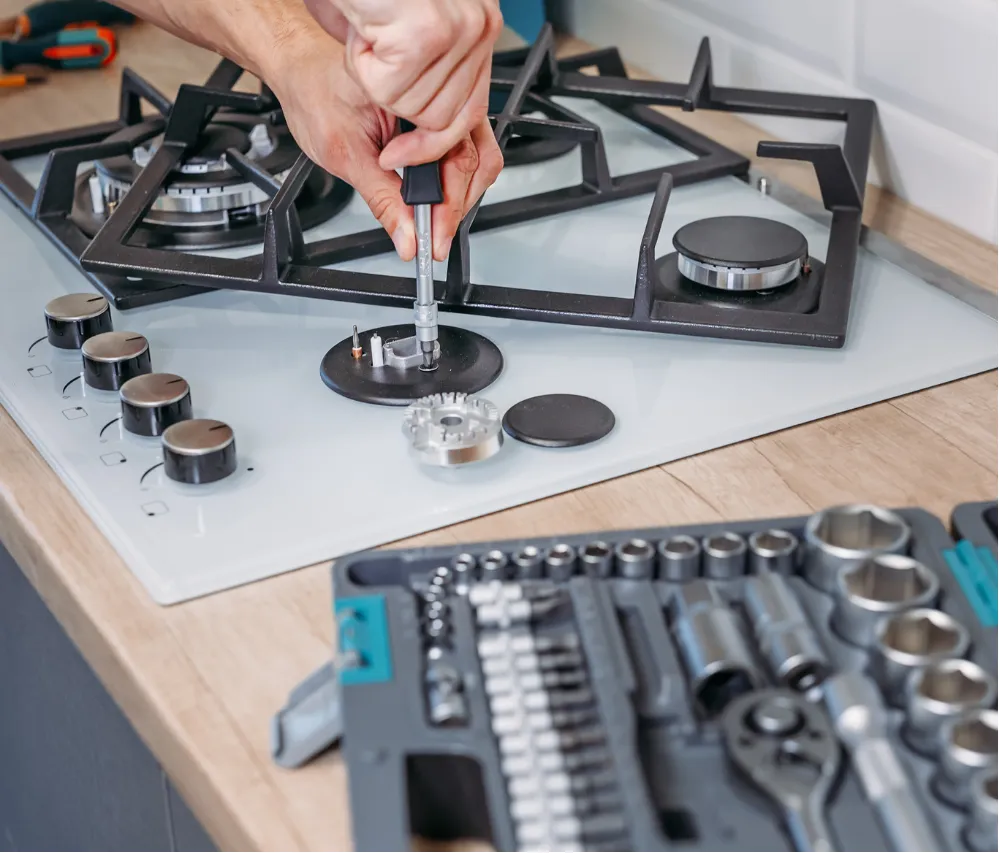 Person using a screwdriver to repair a gas stove with a toolkit on the countertop nearby.