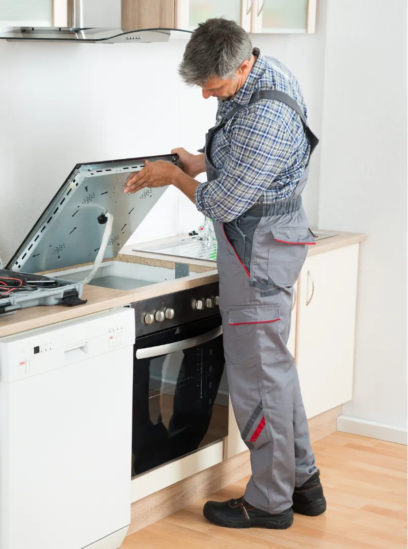 Repairman in overalls fixing an open electric stove in a modern kitchen.