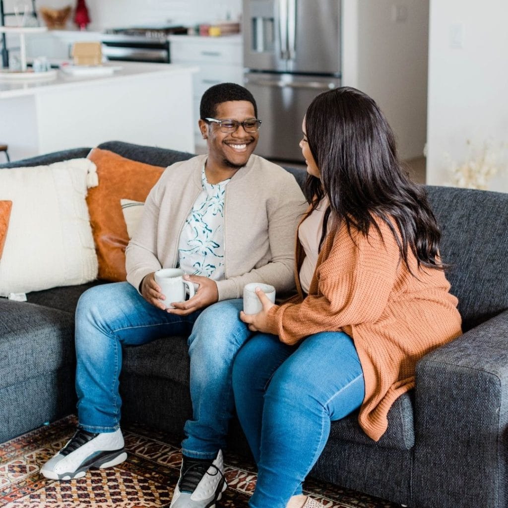 Two people sit on a couch, smiling at each other and holding mugs in a cozy, modern living room.