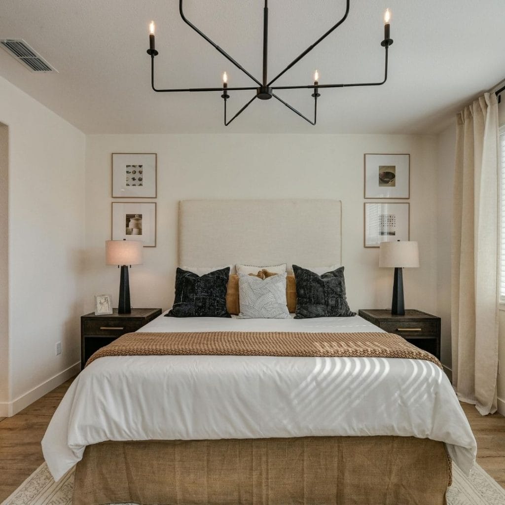 Modern bedroom with a neutral palette, large bed, dark lamps, wall art, and a black geometric chandelier.