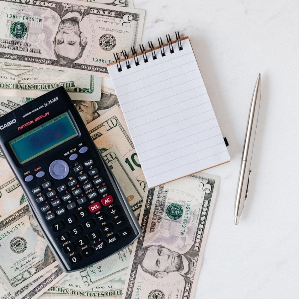 A calculator, pen, and notepad placed on scattered US dollar bills on a white surface.