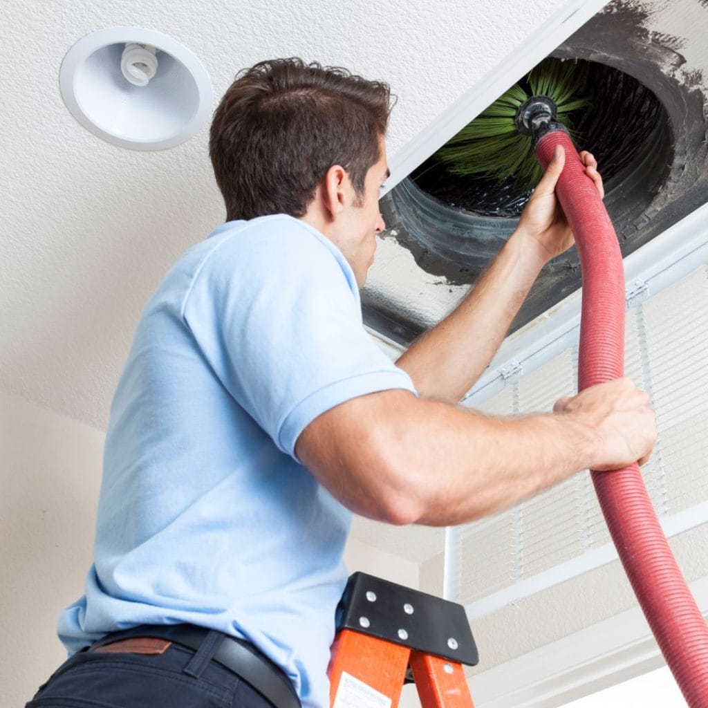 Man standing on ladder cleaning air duct with a vacuum hose inside a ceiling vent.