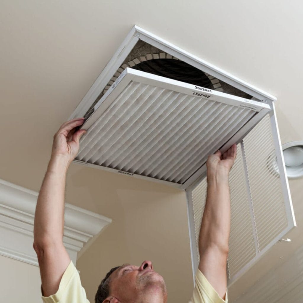 A person changes an air filter in a ceiling vent, reaching up while holding the vent cover open.