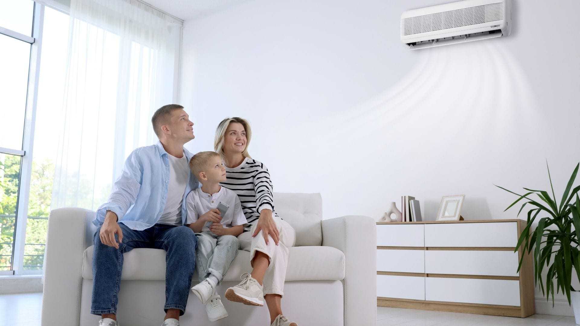 A family of three sits on a couch, smiling and looking at an air conditioner on the wall in a bright living room.