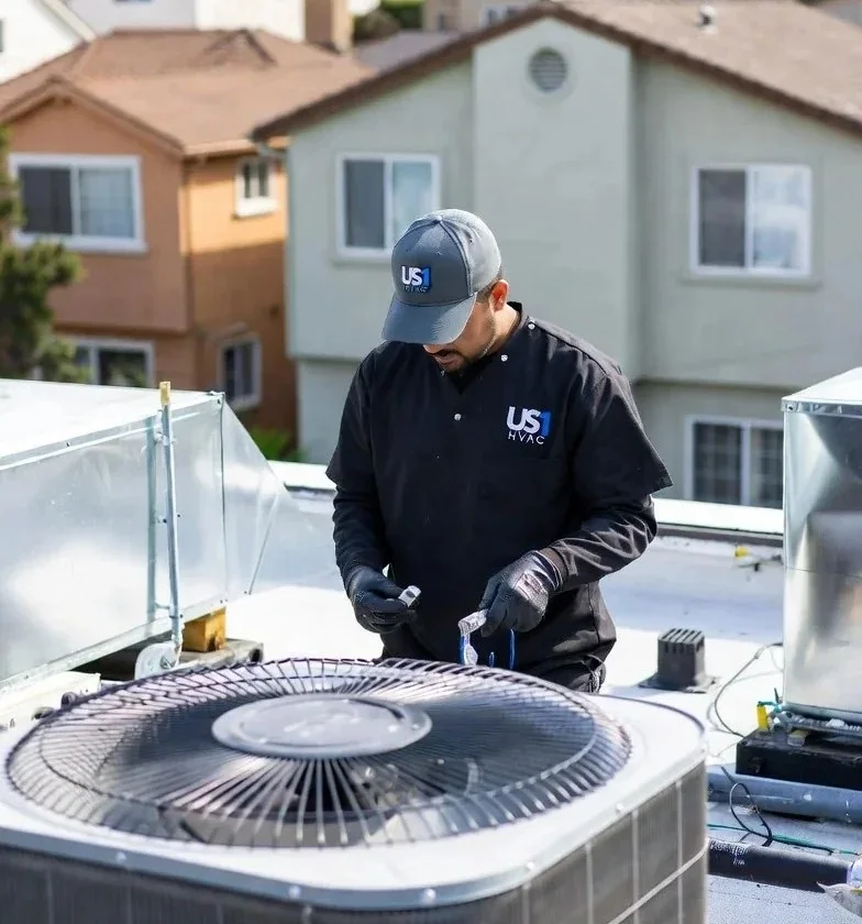 A technician in a US HVAC uniform services an outdoor air conditioning unit on a rooftop.