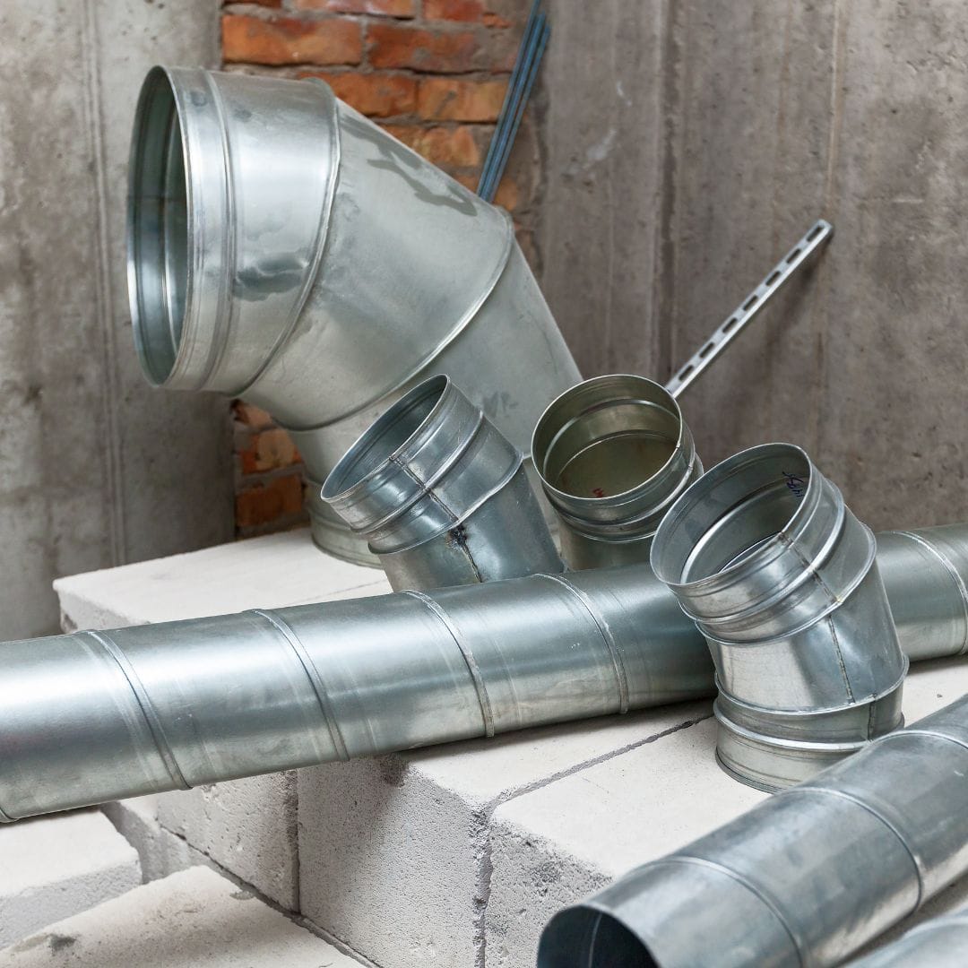 Several metal duct pipes and fittings are placed on concrete blocks against a brick and concrete wall.