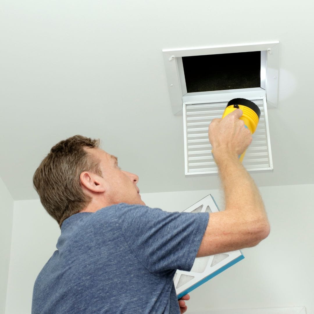 Man inspecting an open ceiling air vent with a flashlight, holding an air filter in his other hand.