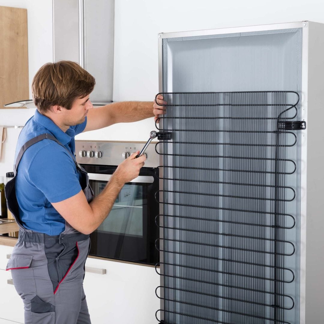 Man in overalls repairing the back of a refrigerator with a screwdriver in a modern kitchen.
