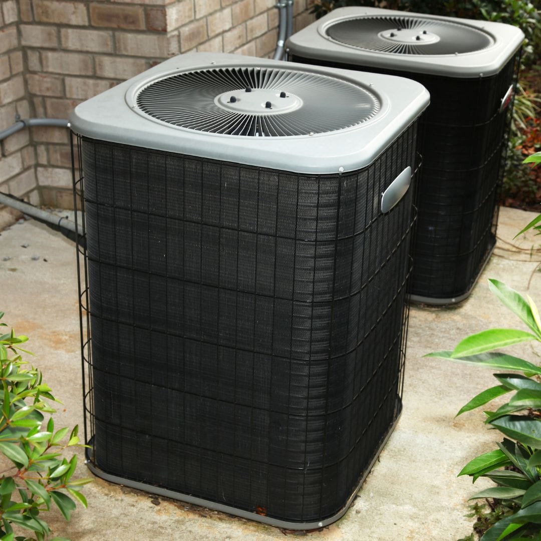 Two black outdoor air conditioning units sit on a concrete pad next to a brick wall and green plants.