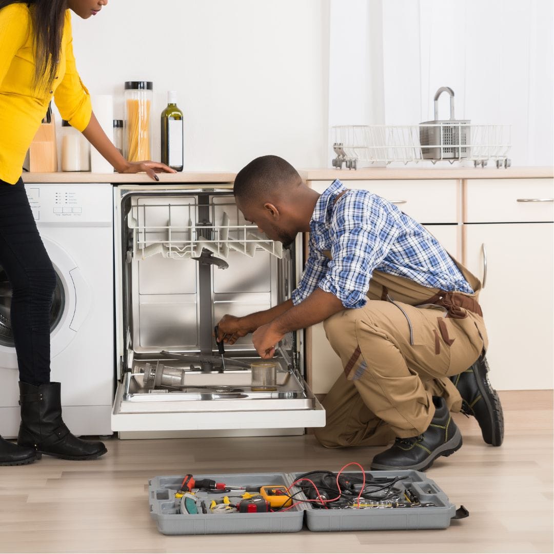 Man repairing a dishwasher with tools while a woman watches in a modern kitchen.