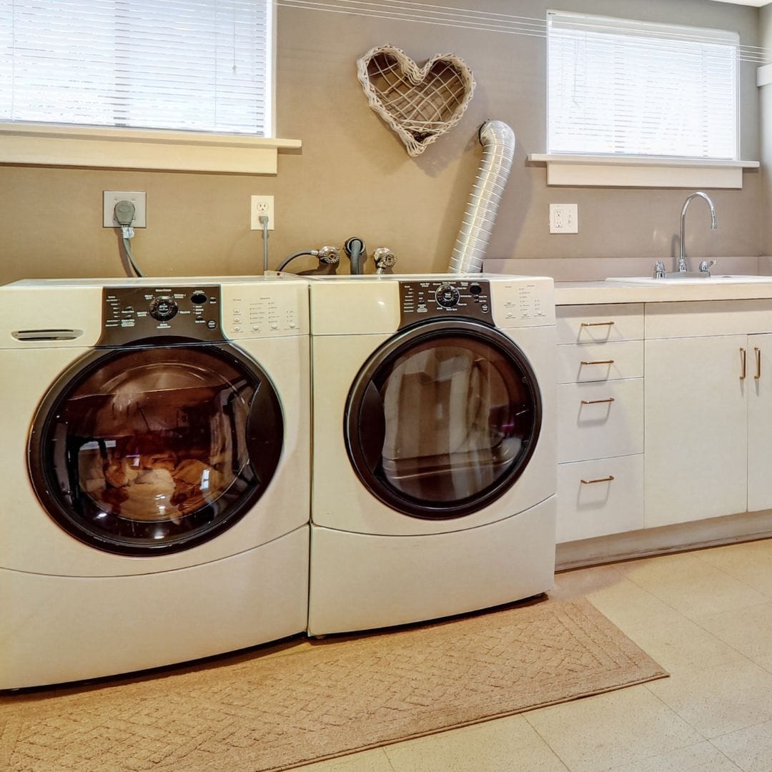 Front-load washer and dryer in a laundry room with beige walls and cabinets, and a heart-shaped wall decor.