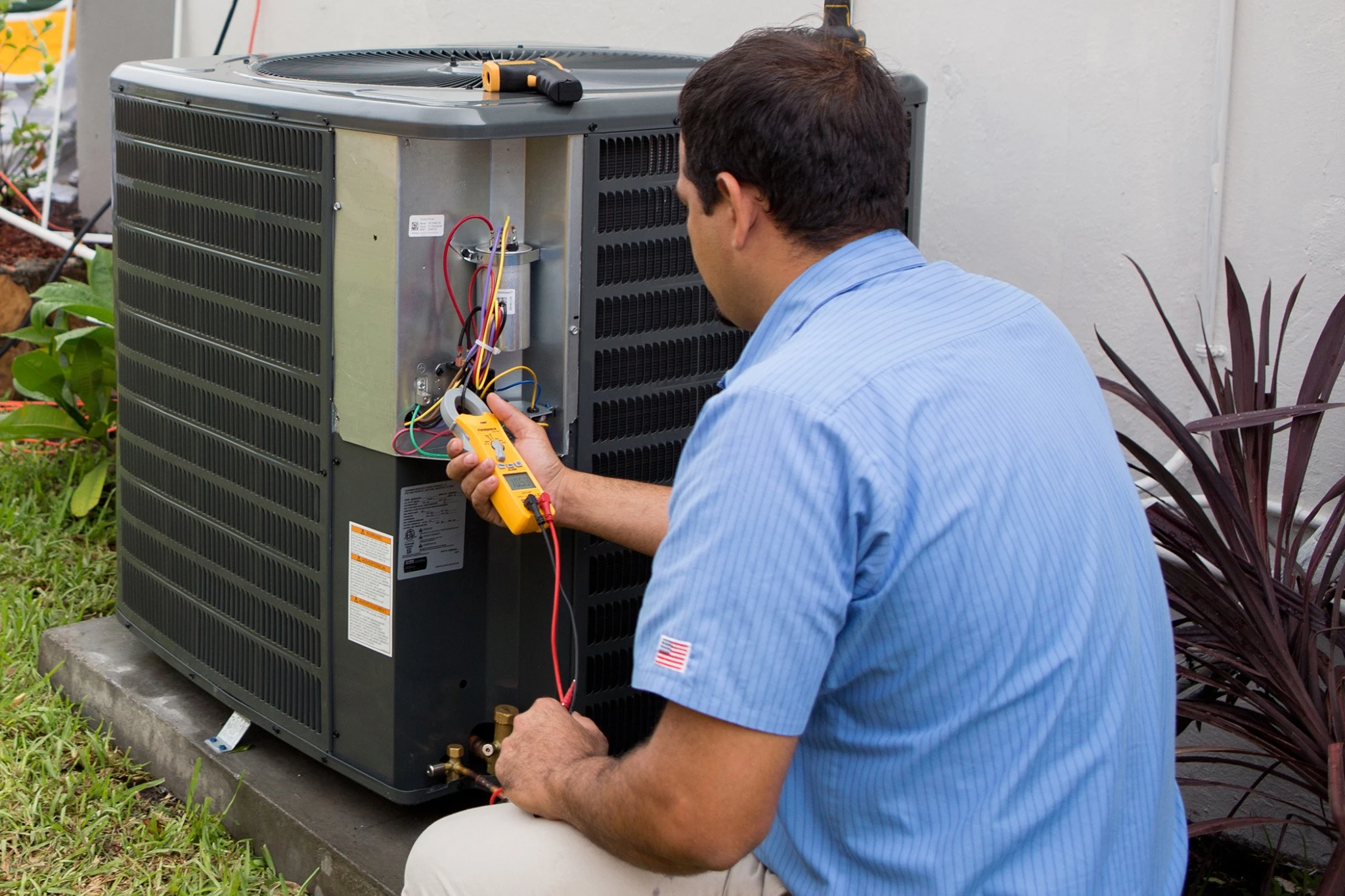 A technician checks an outdoor air conditioning unit with a multimeter near a house.