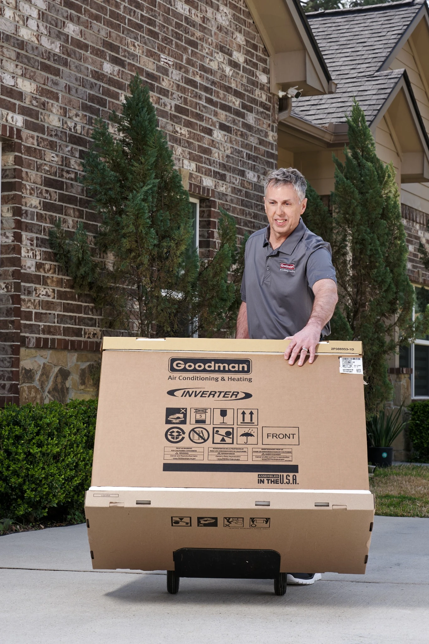 Smiling man moves a large Goodman air conditioner box on a dolly outside a brick house.