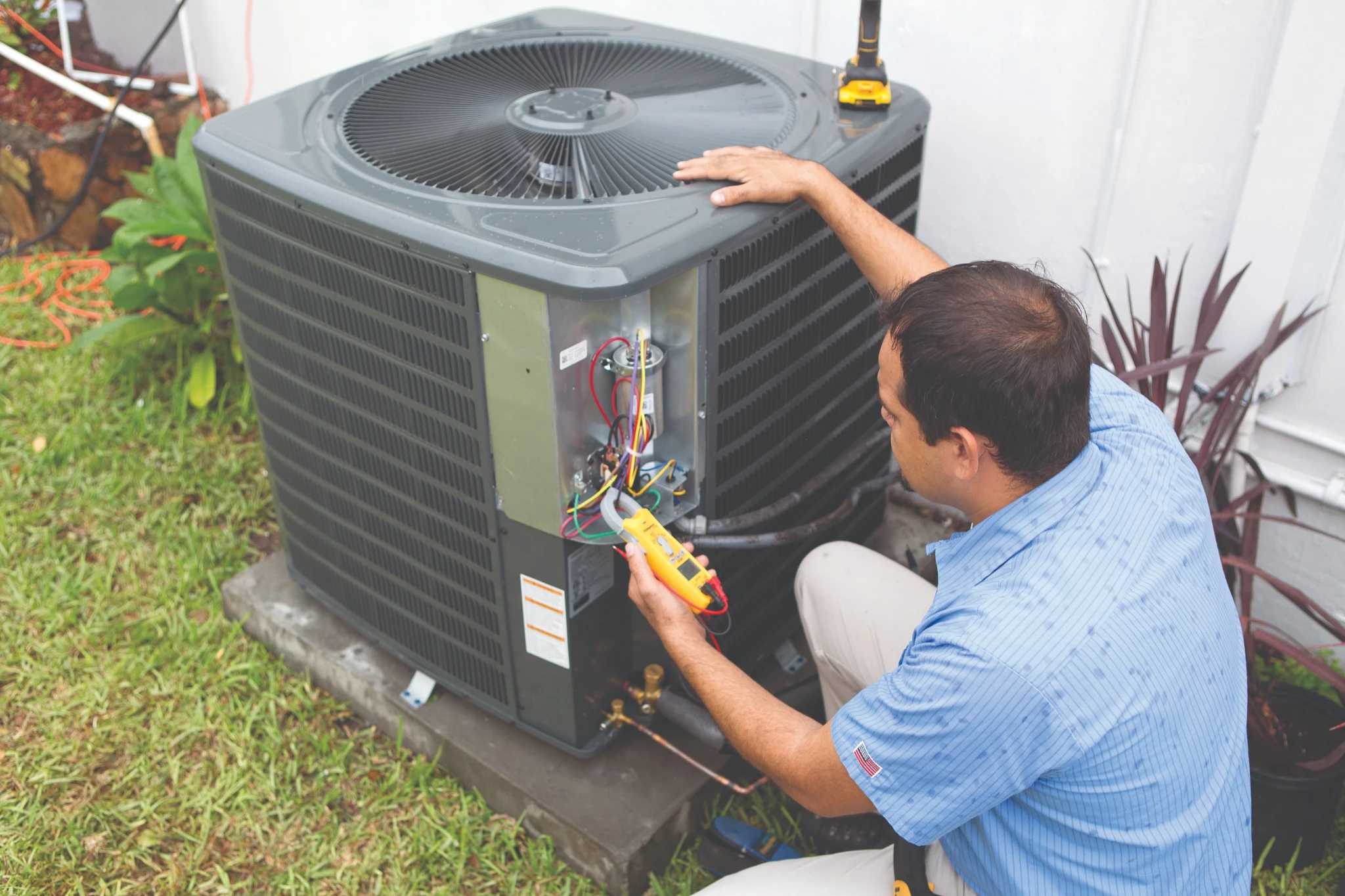 Technician in blue shirt checking wires on an outdoor air conditioning unit with a multimeter.