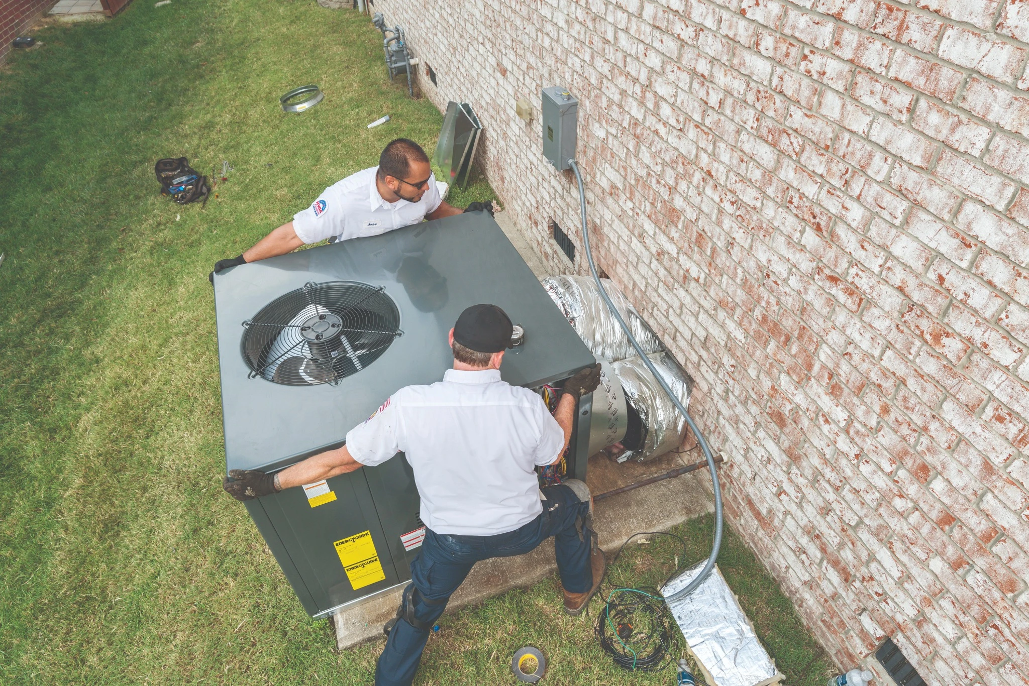 Two technicians install or repair an outdoor air conditioning unit next to a brick building.