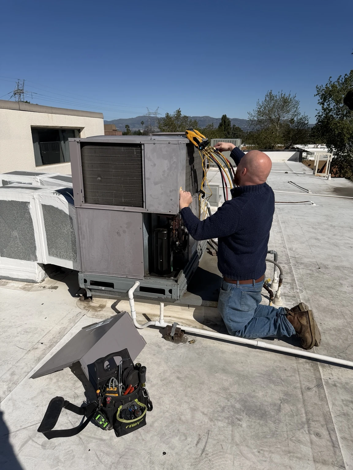 A technician kneels on a rooftop servicing an HVAC unit with tools and gauges in hand.